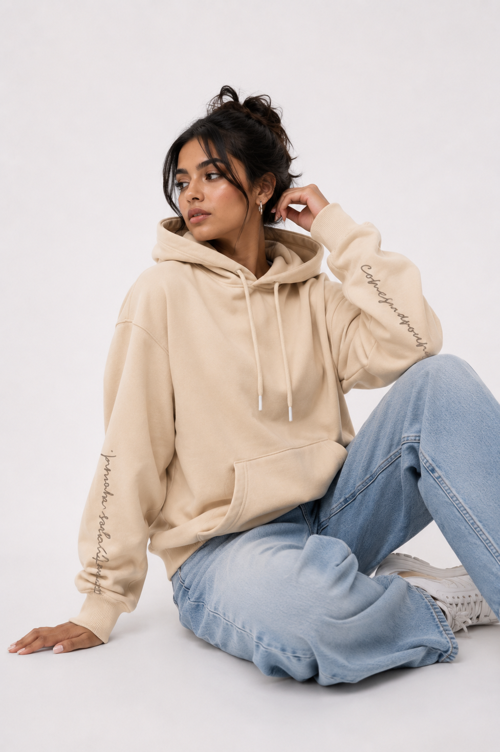 Female model wearing the Karmic Bloom oversized sand beige hoodie in a relaxed seated pose highlighting the oversized fit and sleeve typography.