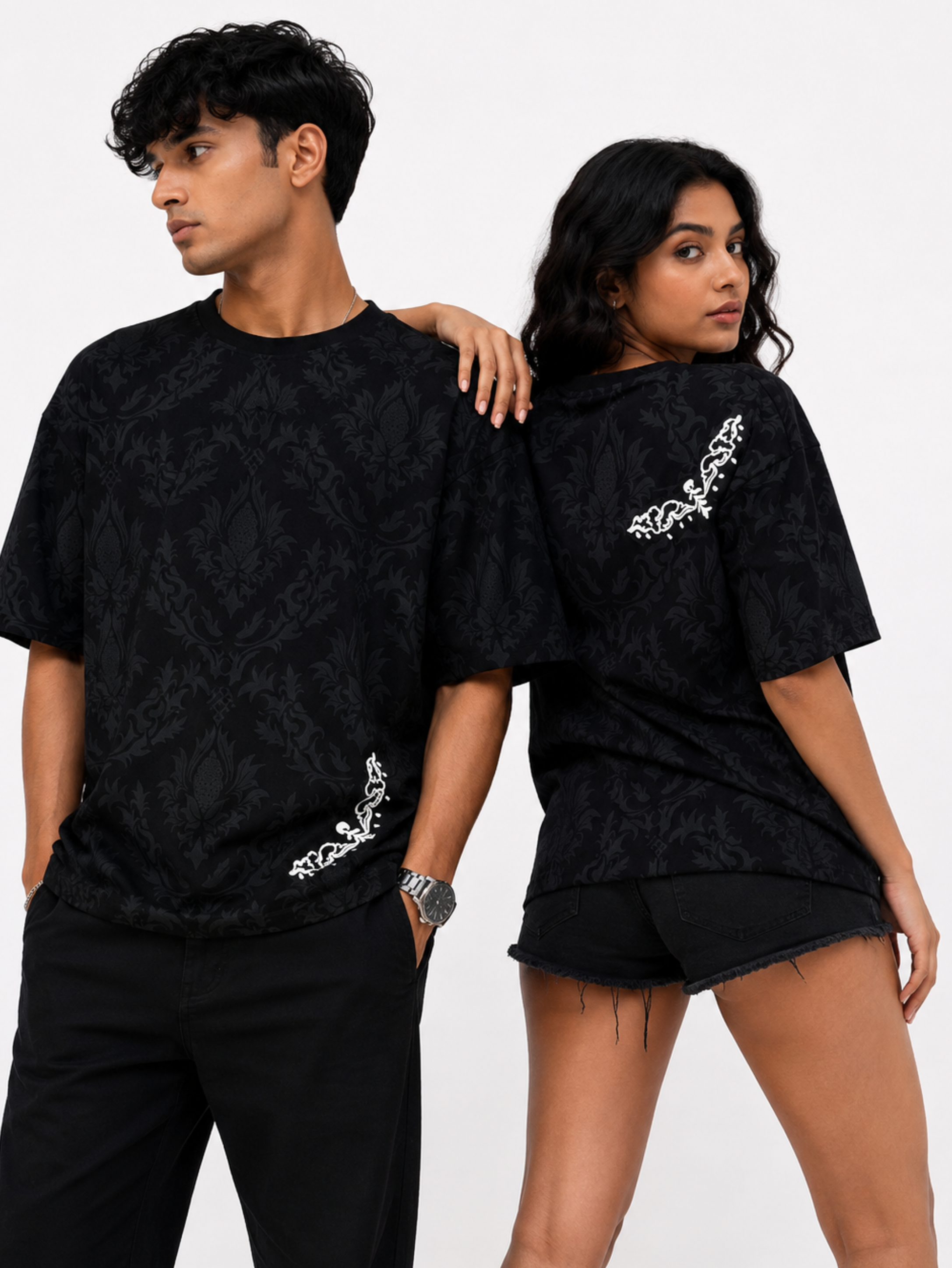 Male and female models wearing the Shadow Moth oversized black heavyweight cotton T-shirt by SYIN & SYRAH featuring a subtle black damask pattern with minimalist white moth graphics on the front and back in a premium studio photoshoot.