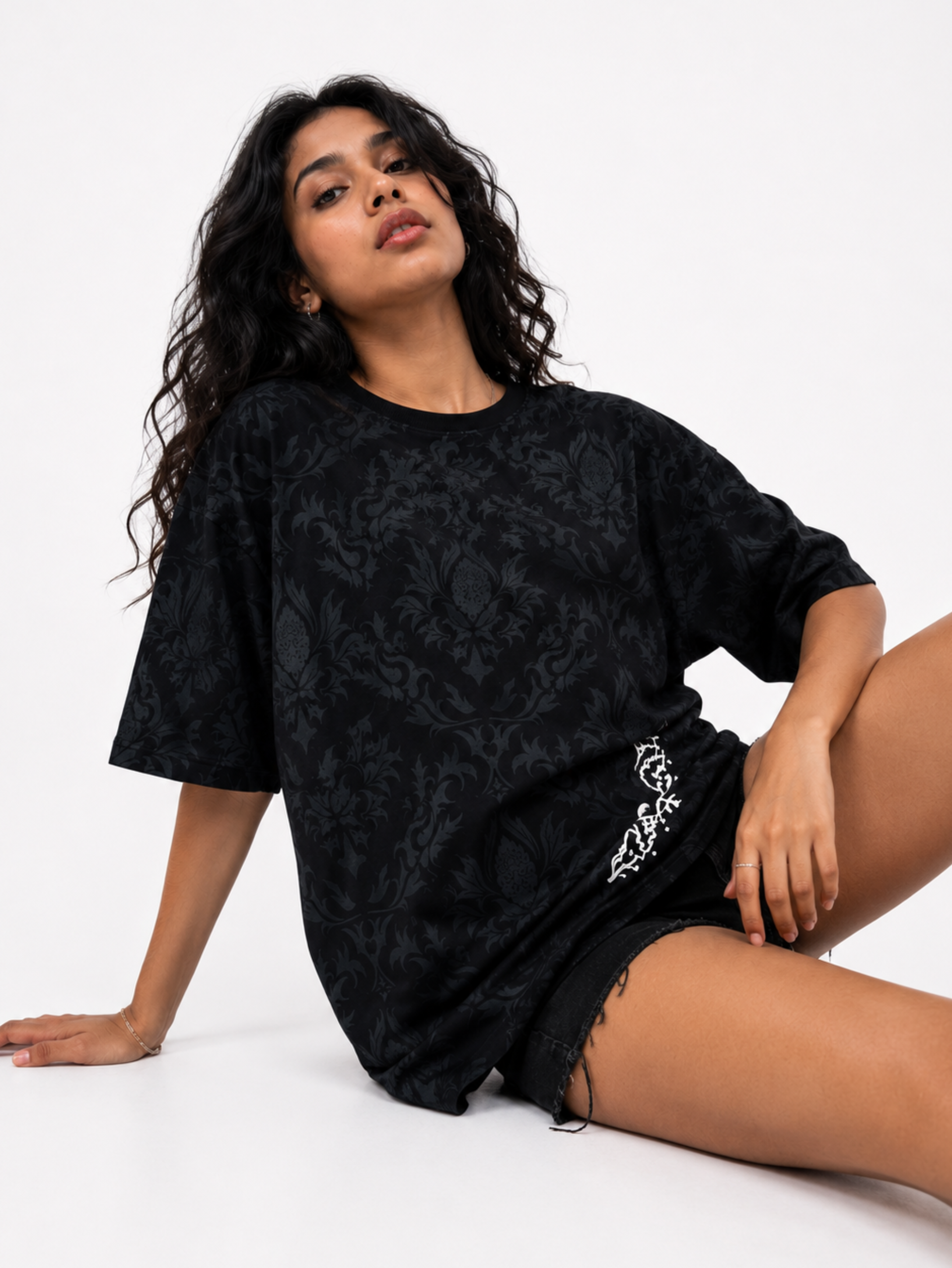 Female model wearing the Shadow Moth oversized black streetwear T-shirt by SYIN & SYRAH featuring an all-over black damask pattern and minimalist white moth graphic in a premium fashion studio shoot.