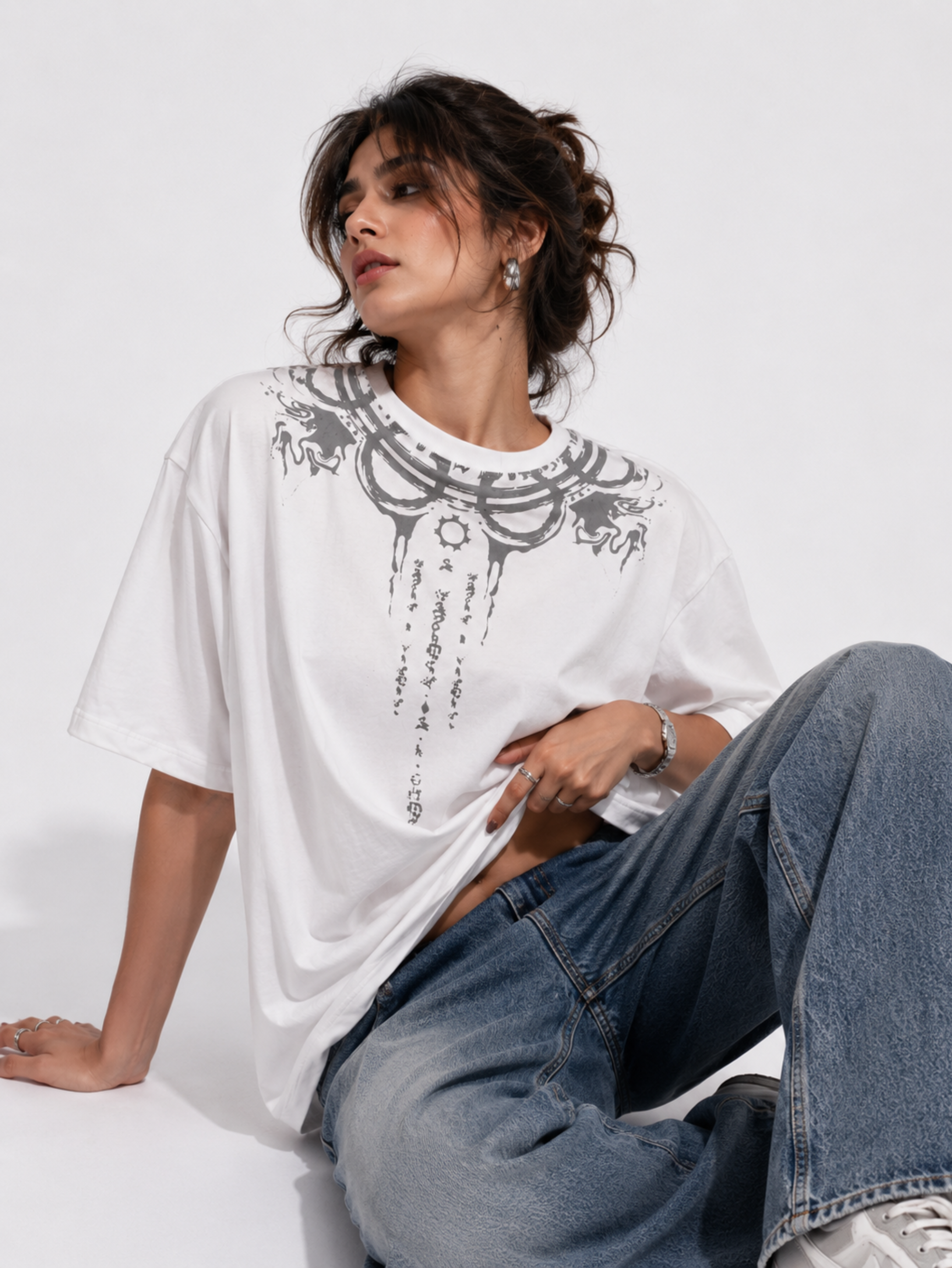 Female model wearing the Vishnu Ascendent premium oversized white cotton T-shirt by Syin&Syrah featuring celestial gothic artwork paired with light blue denim jeans in a modern studio fashion photoshoot.