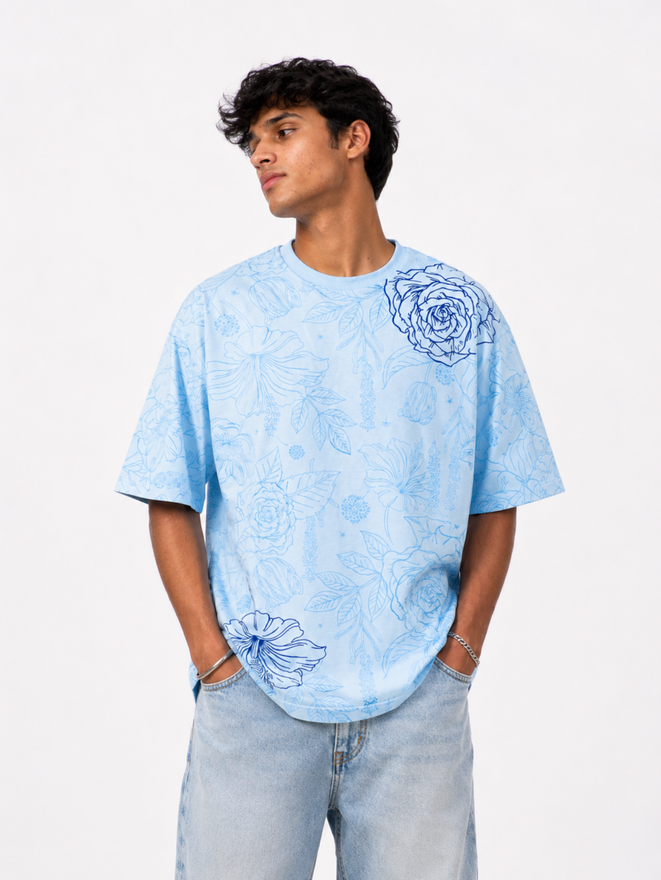 Male model wearing the Vita in Blu premium oversized sky blue cotton T-shirt by SYIN & SYRAH featuring an all-over floral print with blue rose artwork in a white studio setting.