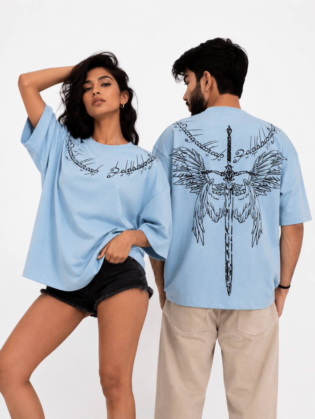Male and female models wearing the Winged Curse oversized sky blue cotton T-shirt by SYIN & SYRAH displaying the front gothic neckline artwork and large winged sword back graphic in a premium studio photoshoot.