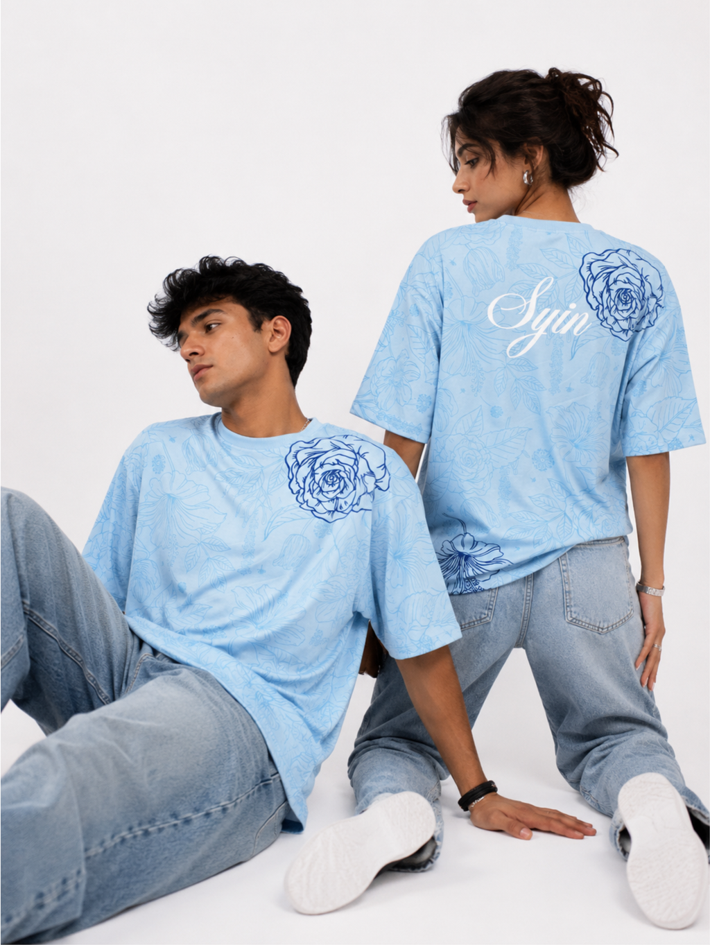 Male and female models wearing the Vita in Blu premium oversized sky blue T-shirt by SYIN & SYRAH displaying the front floral artwork and signature Syin back print in a studio photoshoot.