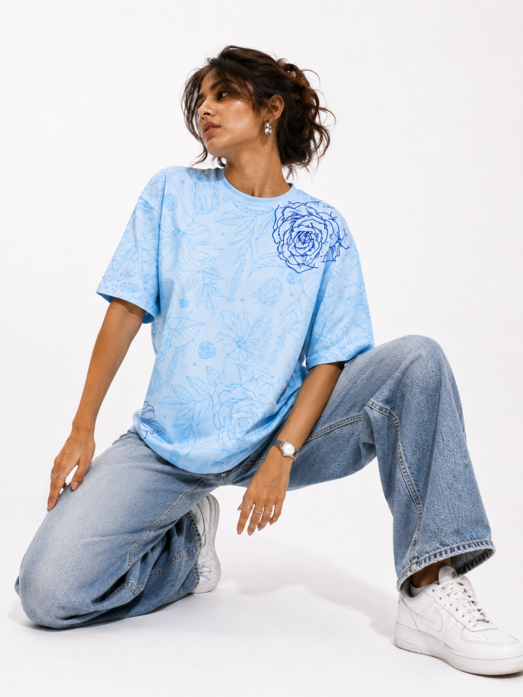 Female model wearing the Vita in Blu premium oversized sky blue floral cotton T-shirt by SYIN & SYRAH paired with light blue denim jeans in a modern studio fashion photoshoot.